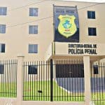 A imagem mostra um prédio bege de quatro andares. Na fachada, há um distintivo amarelo que diz "Polícia Penal". Abaixo, letras pretas dizem "diretoria-geral de polícia penal". Há escadas em frente à porta, e uma grade preta em volta do prédio.