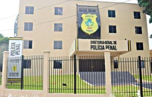 A imagem mostra um prédio bege de quatro andares. Na fachada, há um distintivo amarelo que diz "Polícia Penal". Abaixo, letras pretas dizem "diretoria-geral de polícia penal". Há escadas em frente à porta, e uma grade preta em volta do prédio.