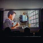Uma tela de cinema se ilumina em frente a um público na penumbra. Uma criança aparece na tela.