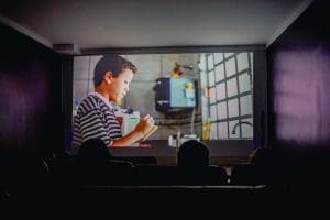 Uma tela de cinema se ilumina em frente a um público na penumbra. Uma criança aparece na tela.