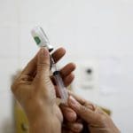 A imagem tem fundo branco e mostra duas mãos brancas preparando uma seringa de vacina.