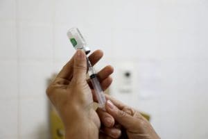 A imagem tem fundo branco e mostra duas mãos brancas preparando uma seringa de vacina.