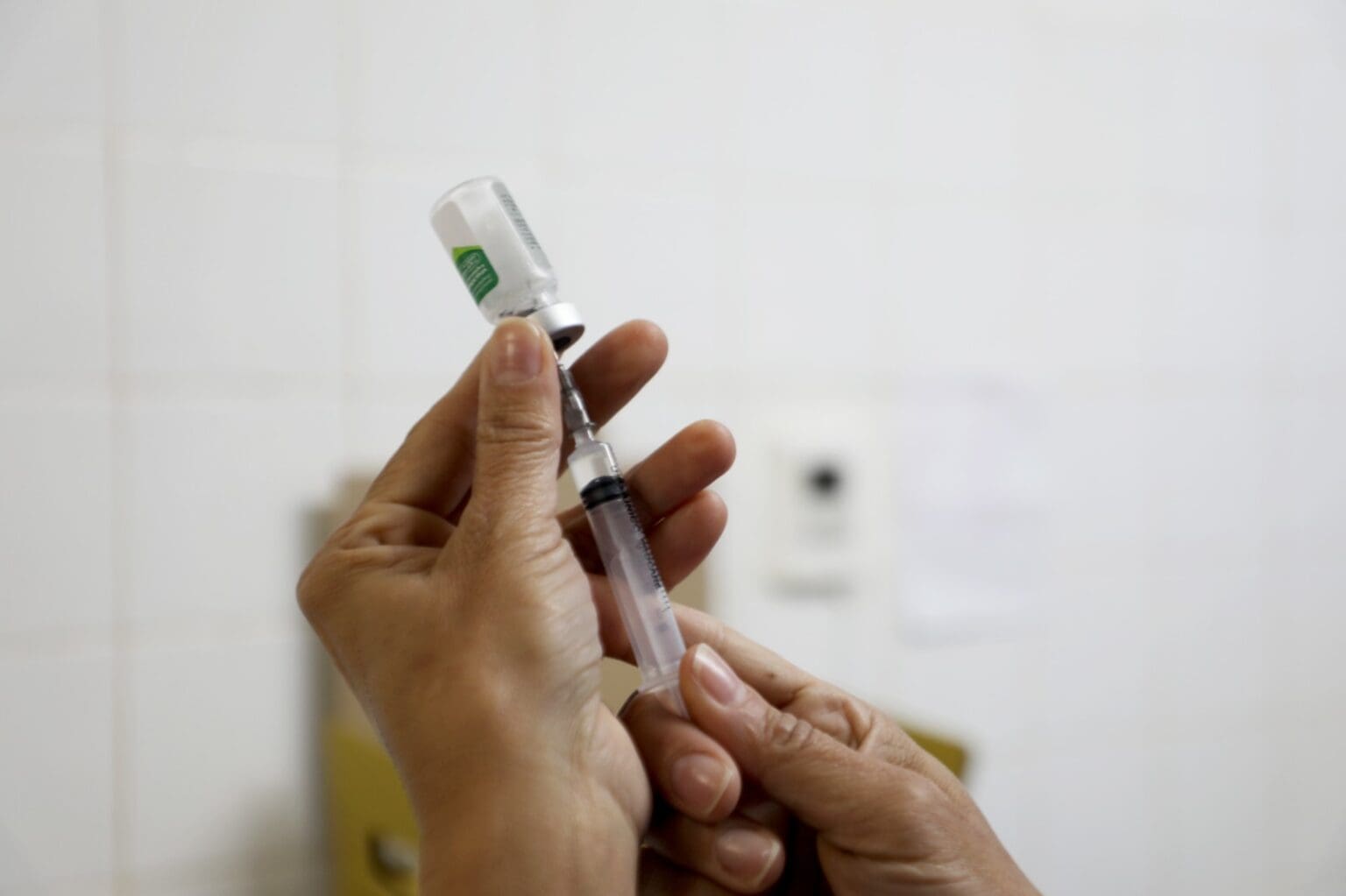 A imagem tem fundo branco e mostra duas mãos brancas preparando uma seringa de vacina.