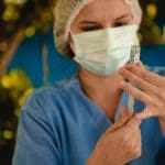 A imagem mostra uma mulher de roupa azul, touca transparente e máscara branca de saúde. Ela está ao fundo e aparece desfocada. A mulher tem uma seringa na mão e prepara uma vacina, que está em primeiro plano.