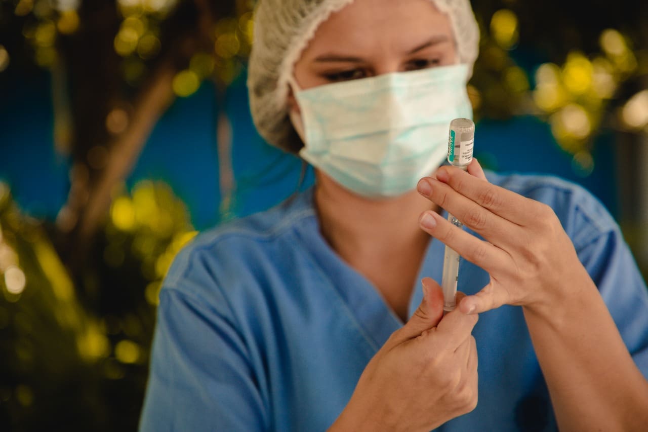 A imagem mostra uma mulher de roupa azul, touca transparente e máscara branca de saúde. Ela está ao fundo e aparece desfocada. A mulher tem uma seringa na mão e prepara uma vacina, que está em primeiro plano.