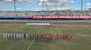 Jogadores de dois times estão enfileirados no meio do campo, esperando que o jogo comece. Uma faixa na arquibancada diz "TOR - Torcida Organizada Rubra".