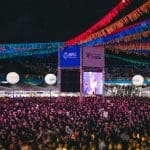 Uma multidão está reunida, o público de um show. Uma torre se ergue no meio, com a logo da Prefeitura de Anápolis e do Voluntários do Coração, com uma tela que mostra o que acontece no palco. Bandeirinhas coloridas estão penduradas sobre o público, amarradas nessa torre.