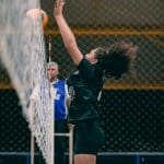 Uma mulher está pulando em frente a uma rede de vôlei, para sacar uma bola. É possível ver o juiz ao fundo.