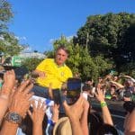 Bolsonaro é um homem branco com cabelos grisalhos. Ele está em cima de um carro, e várias mãos o cumprimentam do lado de fora.
