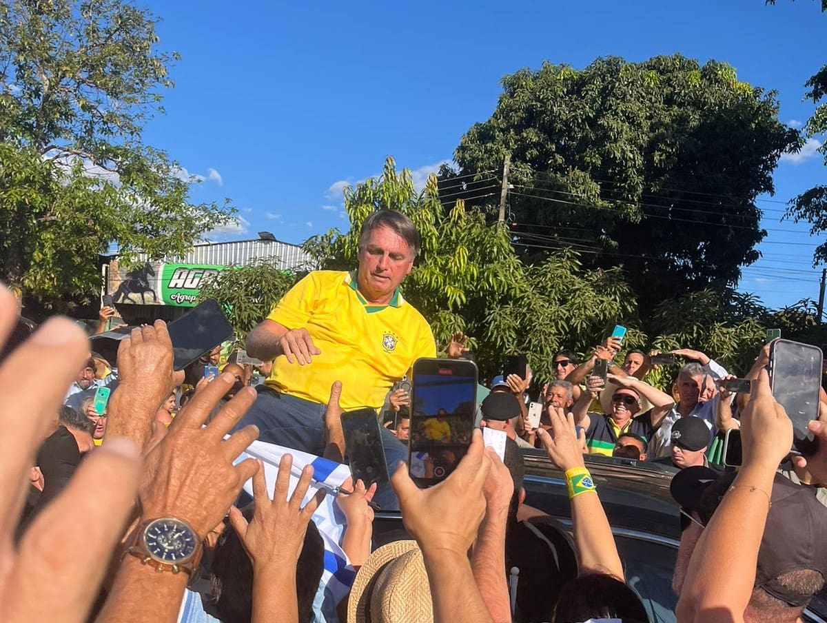 Bolsonaro é um homem branco com cabelos grisalhos. Ele está em cima de um carro, e várias mãos o cumprimentam do lado de fora.