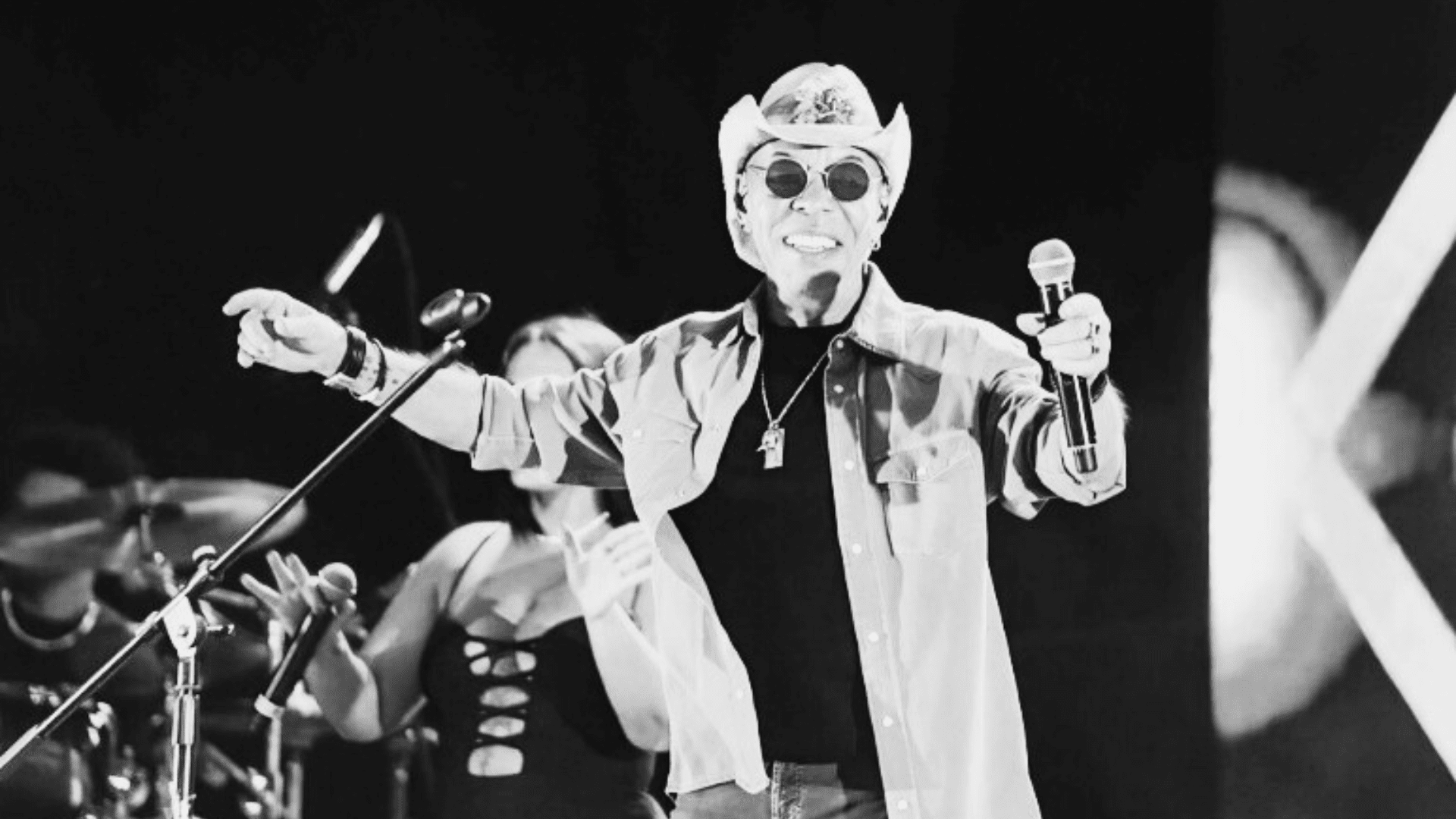 Um homem usa chapéu e óculos escuros. Ele usa uma camisa por cima da camiseta. Tem um microfone na mão e aponta para a plateia.