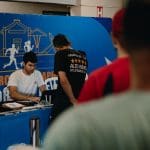 A foto é vista por trás de duas pessoas em uma fila. Mais à frente, um homem sentado faz a inscrição de alguém que está de pé em frente a ele. No fundo, um painel que diz "12º circuito de corrida de rua".