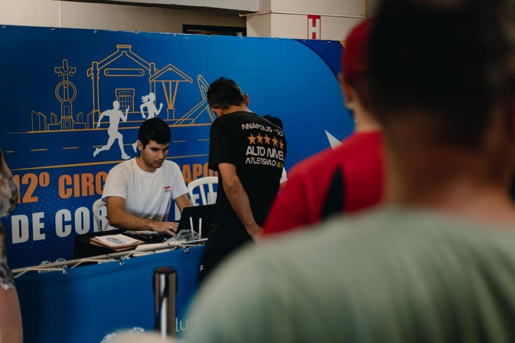 A foto é vista por trás de duas pessoas em uma fila. Mais à frente, um homem sentado faz a inscrição de alguém que está de pé em frente a ele. No fundo, um painel que diz "12º circuito de corrida de rua".