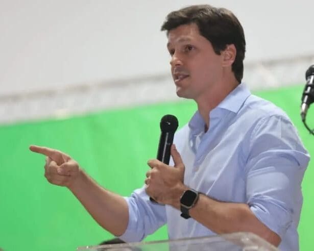 Daniel Vilela é um homem adulto branco de cabelos pretos. Ele usa uma camisa social azul, segura um microfone e aponta para o público enquanto fala.