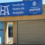 Há uma casa azul claro com portão dourado. A placa acima do portão é azul escuro e diz "ETA - Escola de Teatro de Anápolis".