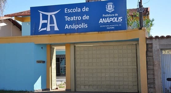 Há uma casa azul claro com portão dourado. A placa acima do portão é azul escuro e diz "ETA - Escola de Teatro de Anápolis".