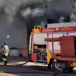 Um galpão cinza pega fogo durante o dia. Um caminhão de bombeiros está em frente.