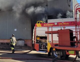 Um galpão cinza pega fogo durante o dia. Um caminhão de bombeiros está em frente.