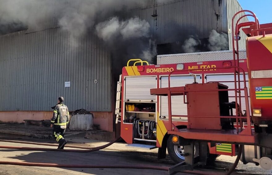 Um galpão cinza pega fogo durante o dia. Um caminhão de bombeiros está em frente.