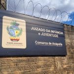 Uma placa em um muro diz: Juizado da infância e juventude, comarca de Anápolis. À esquerda da escrita, um brasão do poder judiciário.