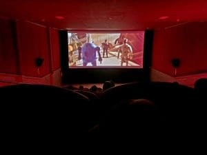 É uma sala de cinema. As luzes estão desligadas, e, na tela, um menino brinca com três bonequinhos. A luz da sala está um pouco avermelhada, e a foto foi tirada do topo da plateia.
