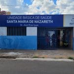 Um posto de saúde é visto pela frente, do outro lado da rua. A fachada azul diz "Unidade básica de saúde Santa Maria de Nazareth, Dr. José Elias Isaac. O portão está aberto e há pessoas lá dentro.