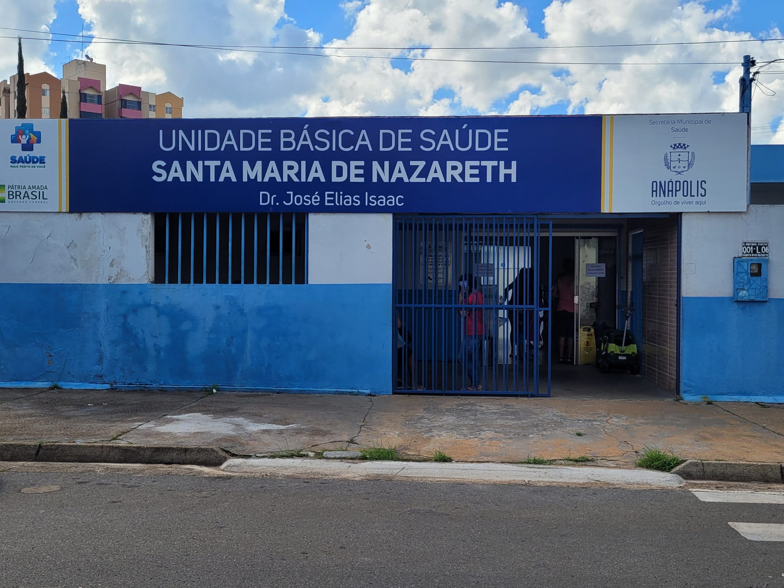 Um posto de saúde é visto pela frente, do outro lado da rua. A fachada azul diz "Unidade básica de saúde Santa Maria de Nazareth, Dr. José Elias Isaac. O portão está aberto e há pessoas lá dentro.