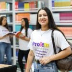 Uma mulher, com cerca de 20 anos, veste uma camiseta branca e sorri para a câmera. Ela tem cabelos escuros longos e lisos. Ao fundo, desfocado, há duas outras mulheres conversando. Elas estão no que parece ser uma biblioteca, pois há estantes de livros ao fundo.