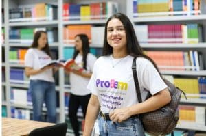 Uma mulher, com cerca de 20 anos, veste uma camiseta branca e sorri para a câmera. Ela tem cabelos escuros longos e lisos. Ao fundo, desfocado, há duas outras mulheres conversando. Elas estão no que parece ser uma biblioteca, pois há estantes de livros ao fundo.