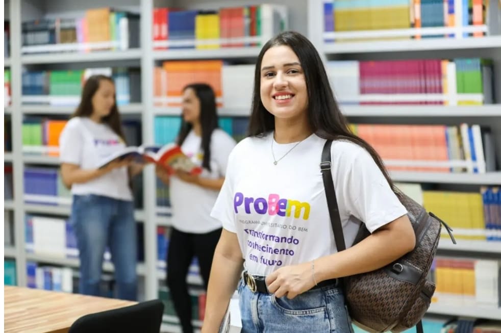 Uma mulher, com cerca de 20 anos, veste uma camiseta branca e sorri para a câmera. Ela tem cabelos escuros longos e lisos. Ao fundo, desfocado, há duas outras mulheres conversando. Elas estão no que parece ser uma biblioteca, pois há estantes de livros ao fundo.
