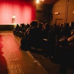 Uma sala com luz avermelhada está cheia de pessoas sentadas em poltronas. À frente da sala, duas pessoas estão em cima de um palco. A porta está à direita, a sala é vista por trás.