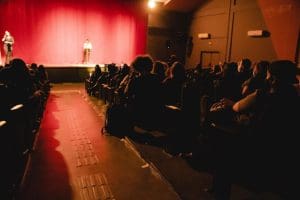 Uma sala com luz avermelhada está cheia de pessoas sentadas em poltronas. À frente da sala, duas pessoas estão em cima de um palco. A porta está à direita, a sala é vista por trás.