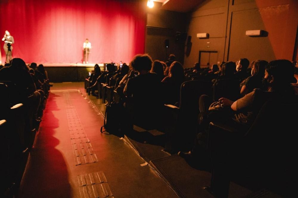 Uma sala com luz avermelhada está cheia de pessoas sentadas em poltronas. À frente da sala, duas pessoas estão em cima de um palco. A porta está à direita, a sala é vista por trás.