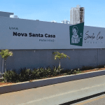 Um prédio branco com as escritas "Uma nova Santa Casa para você". O prédio é da Santa Casa de Anápolis, e uma imagem em verde de São Francisco está ao lado da logo do hospital.