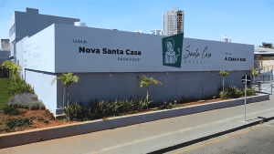 Um prédio branco com as escritas "Uma nova Santa Casa para você". O prédio é da Santa Casa de Anápolis, e uma imagem em verde de São Francisco está ao lado da logo do hospital.