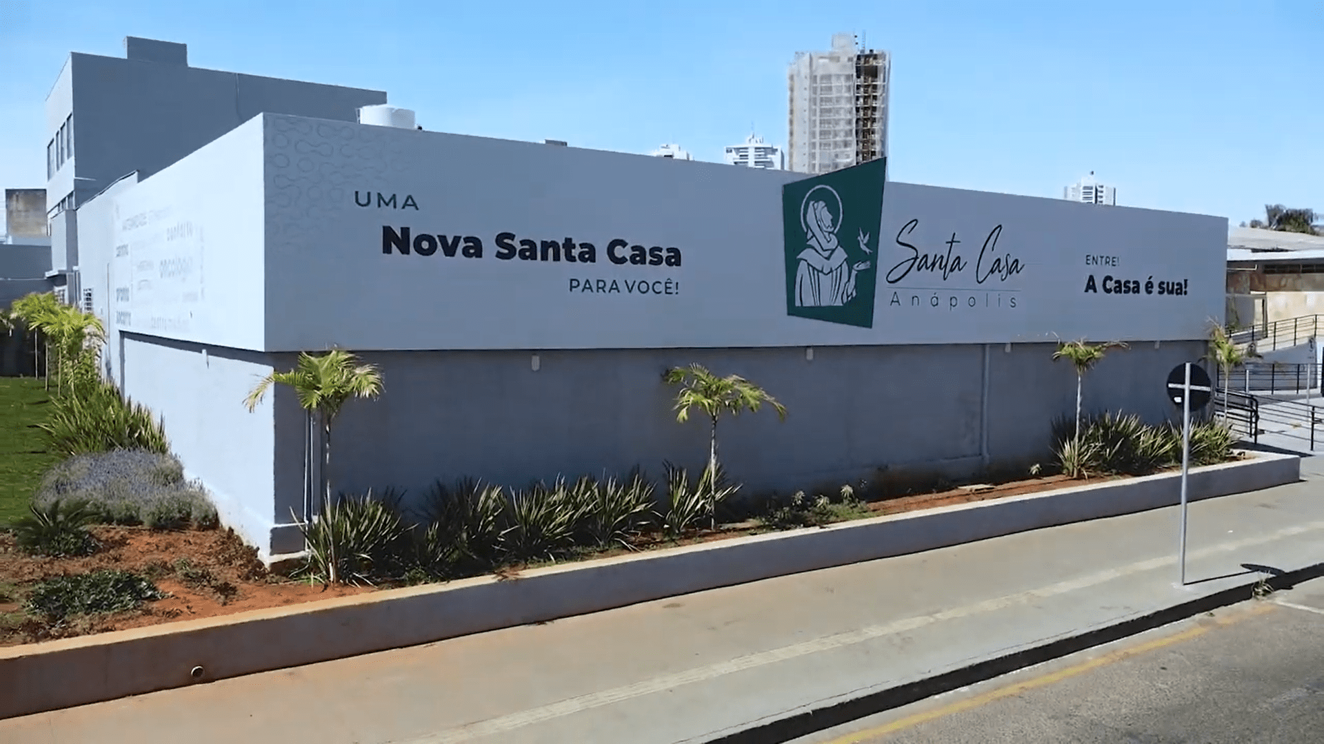 Um prédio branco com as escritas "Uma nova Santa Casa para você". O prédio é da Santa Casa de Anápolis, e uma imagem em verde de São Francisco está ao lado da logo do hospital.