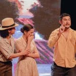 Três pessoas encenam em um palco. A primeira, uma mulher, usa um chapéu amarelo e camisa branca de botões. Ela abraça a segunda, que está chorando. A segunda mulher usa um vestido. A terceira pessoa é um homem branco de cabelos pretos, que aponta os dois dedos indicadores para o céu e fala algo.