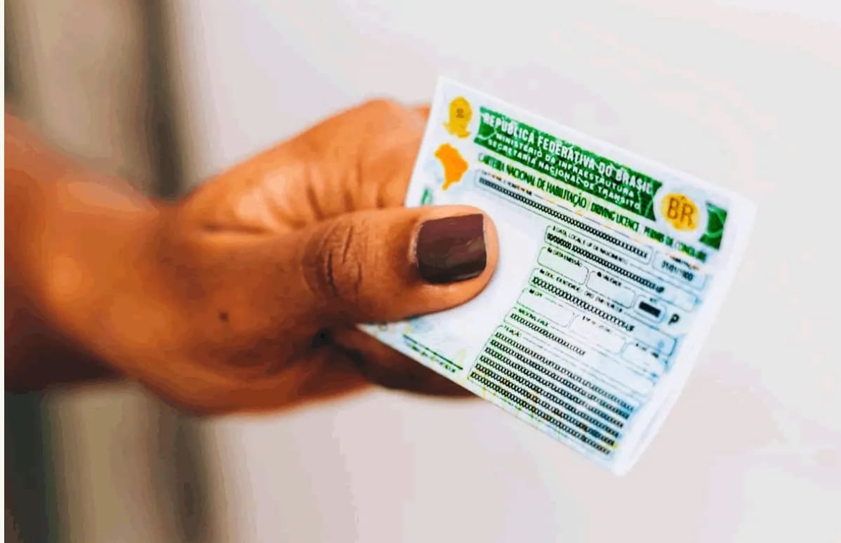 Uma mão com unha pintada segura uma carteira nacional de habilitação, de cor verde. Não há nada no fundo.