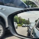 Um retrovisor dianteiro é visto da janela do motorista de um carro. No espelho, o reflexo do carro a que pertence o retrovisor. Ao fundo, é possível ver outro carro passando pela rua.