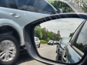 Um retrovisor dianteiro é visto da janela do motorista de um carro. No espelho, o reflexo do carro a que pertence o retrovisor. Ao fundo, é possível ver outro carro passando pela rua.