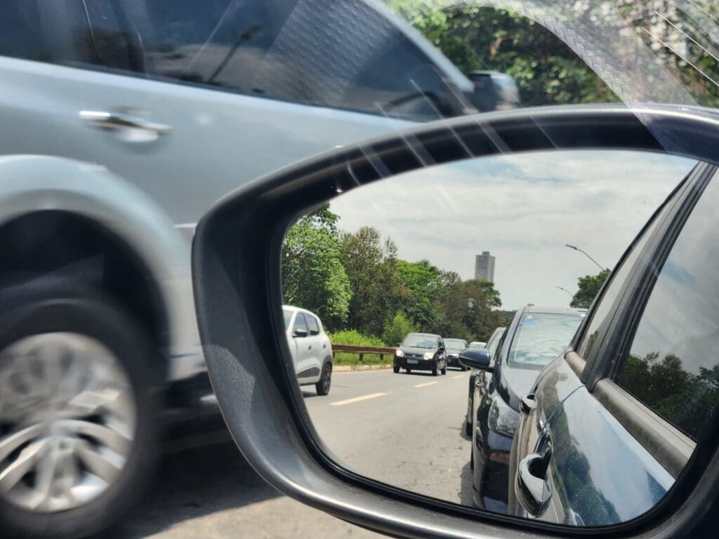 Um retrovisor dianteiro é visto da janela do motorista de um carro. No espelho, o reflexo do carro a que pertence o retrovisor. Ao fundo, é possível ver outro carro passando pela rua.