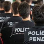 Várias pessoas são vistas pelas costas, sentados em cadeiras enquanto assistem a algo que está fora da tela. Todos têm cabelos curtos escuros e vestem um uniforme preto da polícia penal.