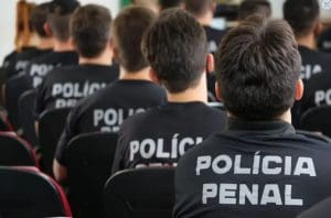 Várias pessoas são vistas pelas costas, sentados em cadeiras enquanto assistem a algo que está fora da tela. Todos têm cabelos curtos escuros e vestem um uniforme preto da polícia penal.