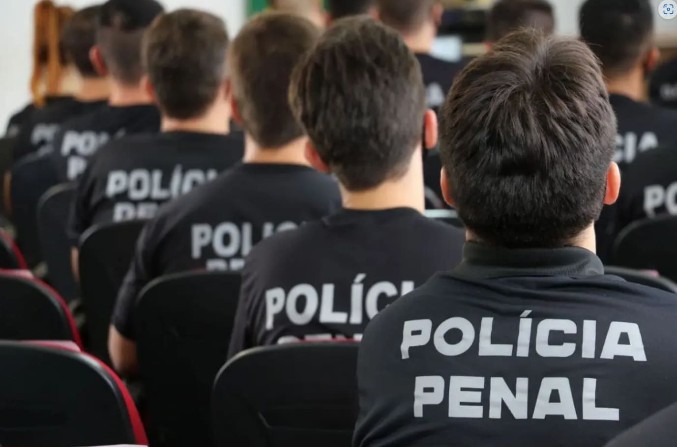 Várias pessoas são vistas pelas costas, sentados em cadeiras enquanto assistem a algo que está fora da tela. Todos têm cabelos curtos escuros e vestem um uniforme preto da polícia penal.