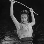 Uma cena de filme em preto e branco. Não há nada ao fundo. Uma mulher japonesa veste roupas tradicionais e levanta uma espada sobre a cabeça.