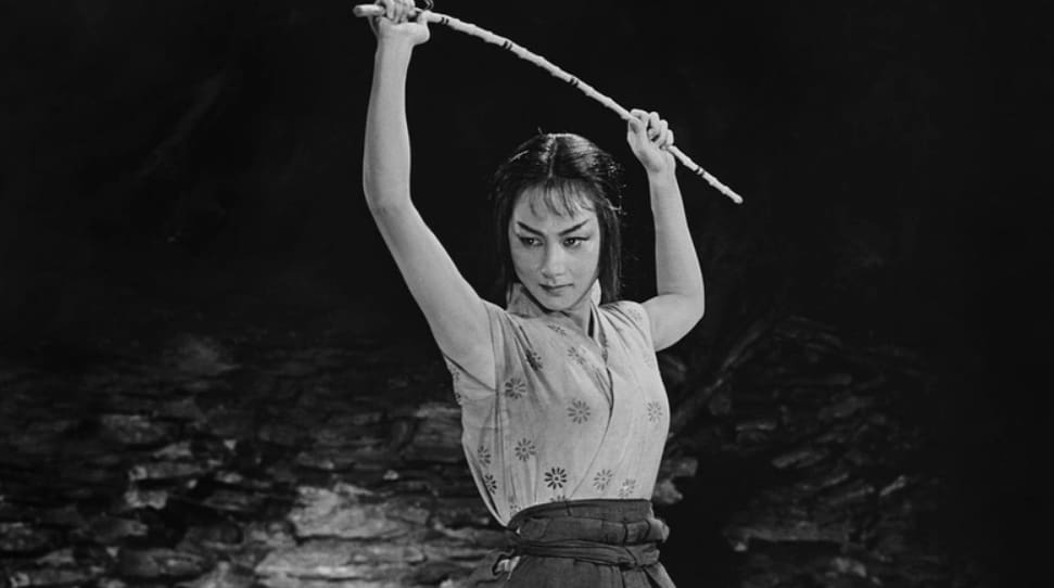 Uma cena de filme em preto e branco. Não há nada ao fundo. Uma mulher japonesa veste roupas tradicionais e levanta uma espada sobre a cabeça.