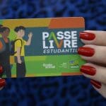 Duas mãos com unhas pintadas de vermelho seguram um cartão escrito Passe Livre. Há a ilustração de dois jovens no cartão colorido.