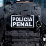 Uma pessoa é vista pelas costas. Ela veste um uniforme totalmente preto e colete. No colete, há as palavras "Polícia penal" em branco.