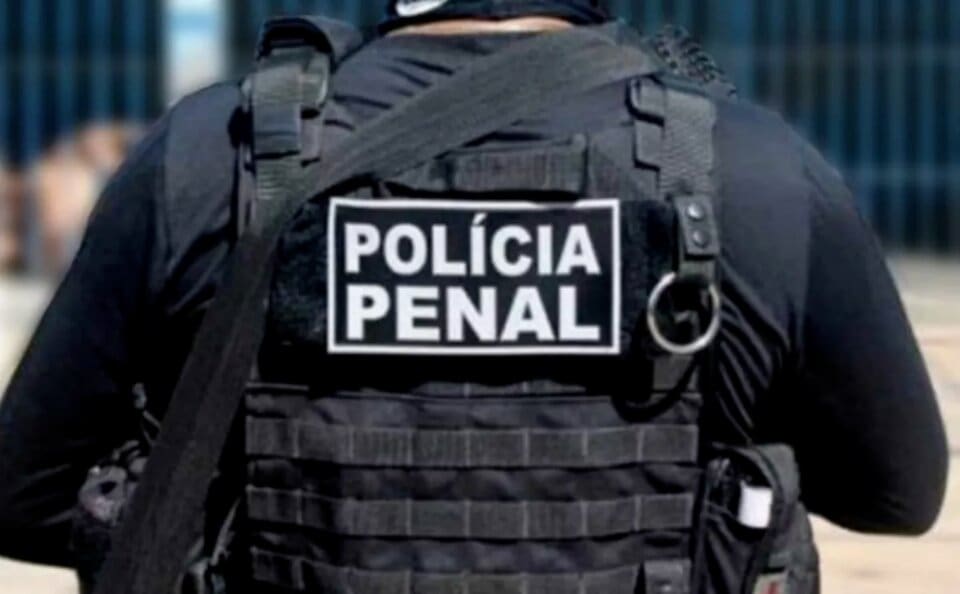 Uma pessoa é vista pelas costas. Ela veste um uniforme totalmente preto e colete. No colete, há as palavras "Polícia penal" em branco.