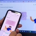 Em plano de fundo, o site do Prouni está aberto em uma tela de computador. Em primeiro plano, duas mãos seguram um celular, onde também está aberto o site do Prouni.